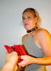  A woman is holding a red wallet over a woman's shoulder and she is demanding money. The woman with the wallet is wearing a black collar and a black shirt.