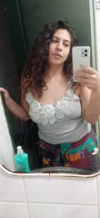 A woman takes a selfie in the mirror wearing a grey tank top and white pants